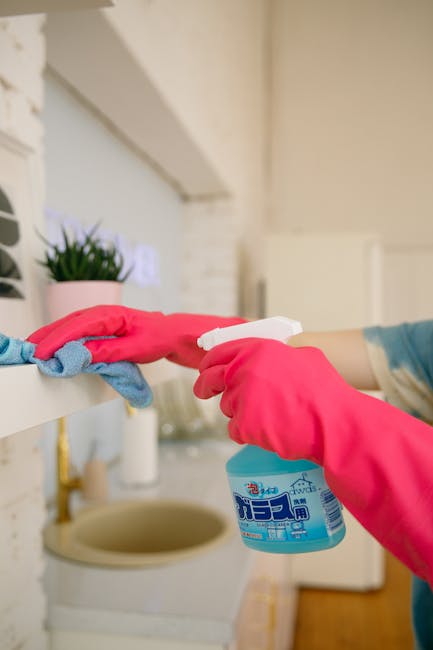 A person wearing bright pink rubber gloves is holding a spray bottle of cleaning solution in one hand and using a cloth to clean a white shelf in a domestic setting. On the shelf, there is a small pot